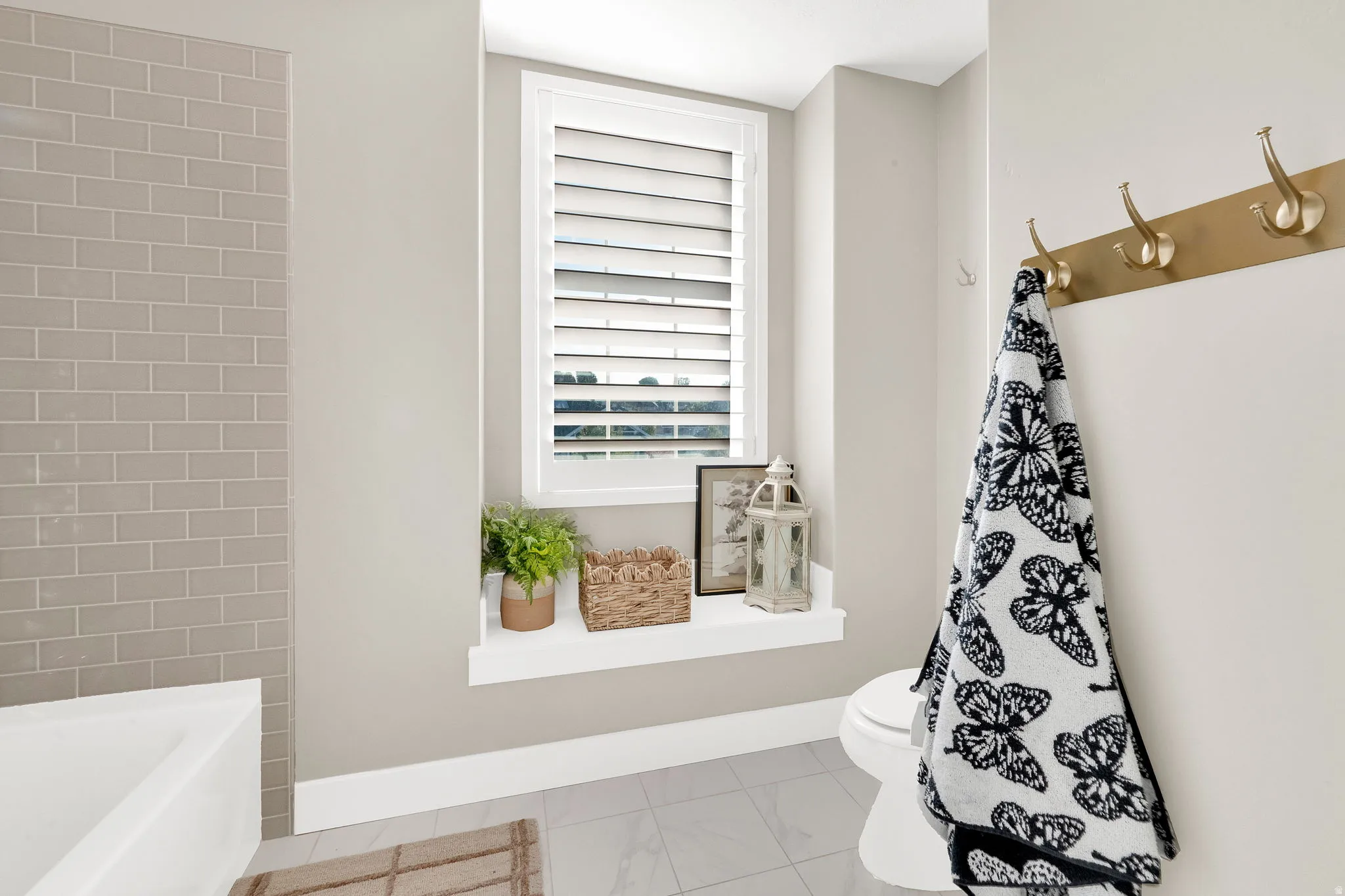 Recessed window with plantation shutters, light gray subway tile shower surround, white baseboards, and wall-mounted brass coat hooks