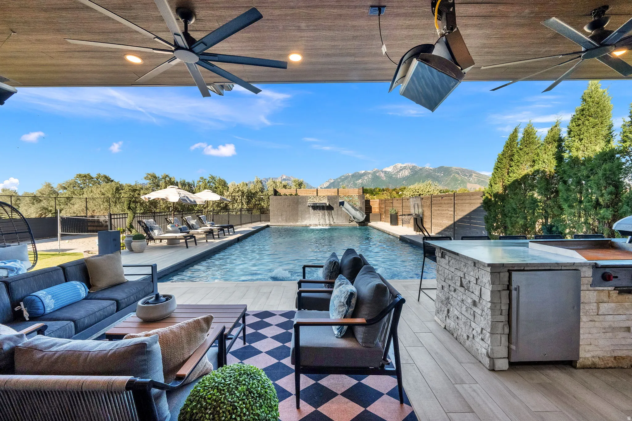 Covered outdoor patio featuring a large swimming pool with water feature, integrated stone outdoor kitchen with built-in grill, and mountain backdrop