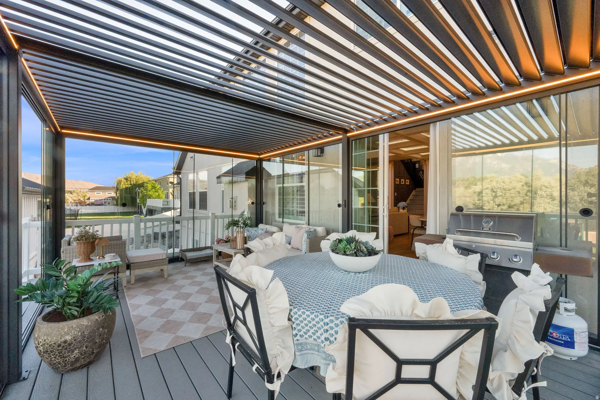 Expansive outdoor living space featuring a louvered pergola with integrated lighting, glass walls, a built-in outdoor grill station, and wood-finish decking