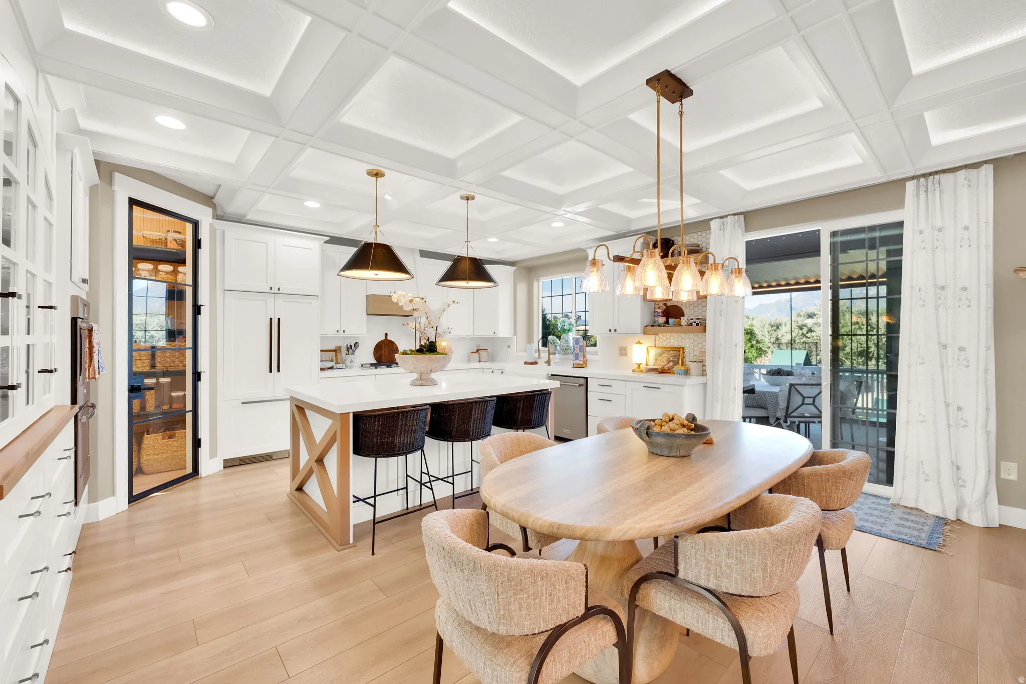 Gourmet kitchen and dining area featuring a coffered ceiling, light wood-finish flooring, white cabinetry, a large kitchen island with seating, and a walk-in pantry with a glass door