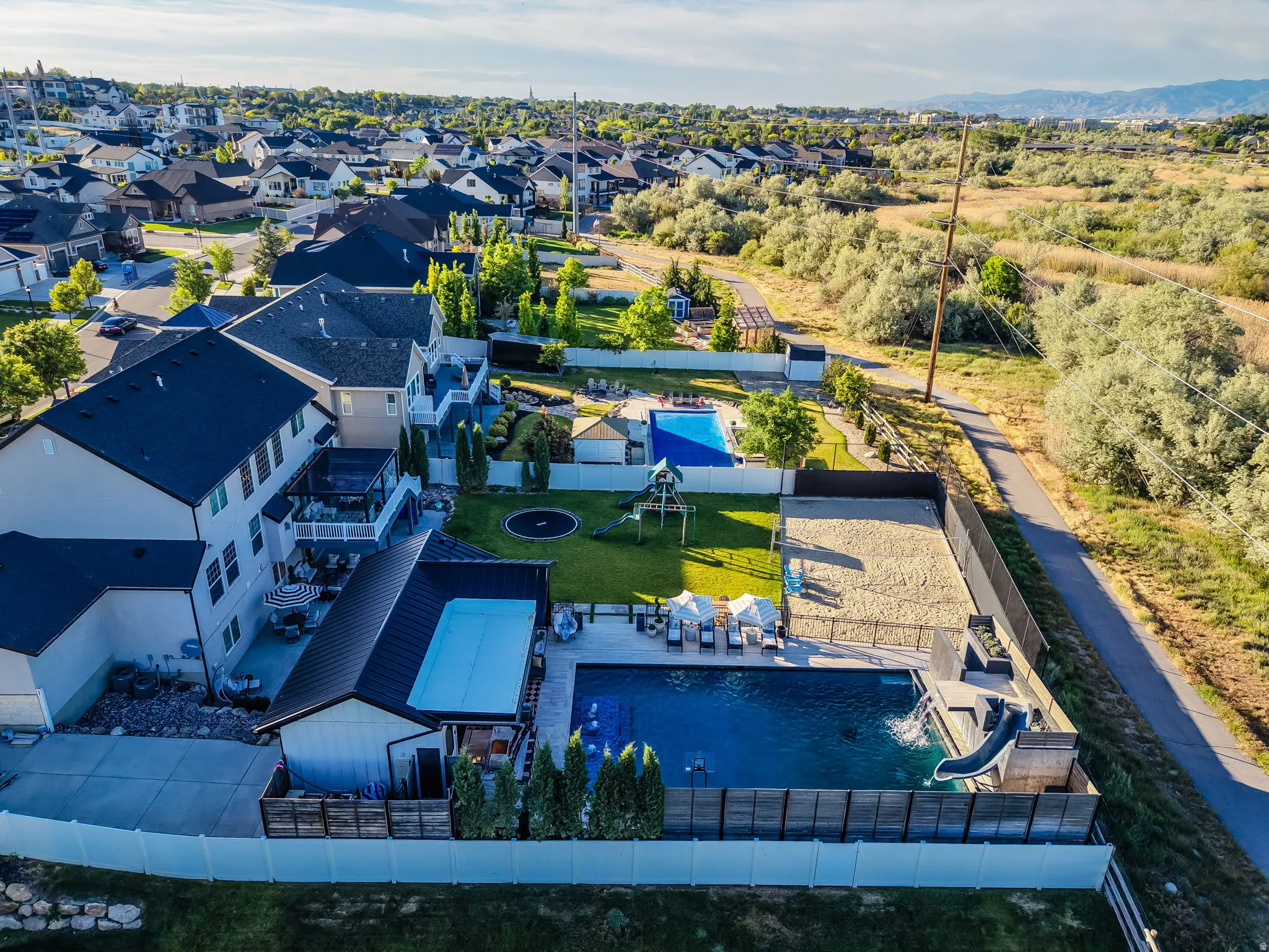 Expansive backyard featuring a large in-ground swimming pool with an attached slide, a dedicated sand court, a separate trampoline area, and a secondary pool