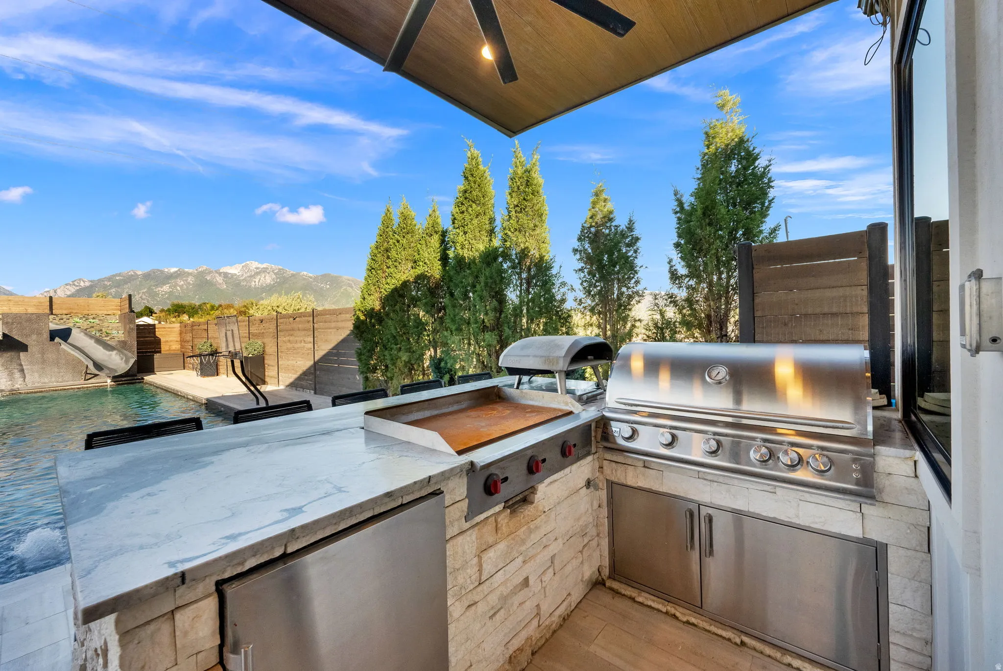 Outdoor kitchen featuring a built-in stainless steel grill, a flat-top griddle, integrated refrigeration, and a stone-clad base with a light-toned countertop