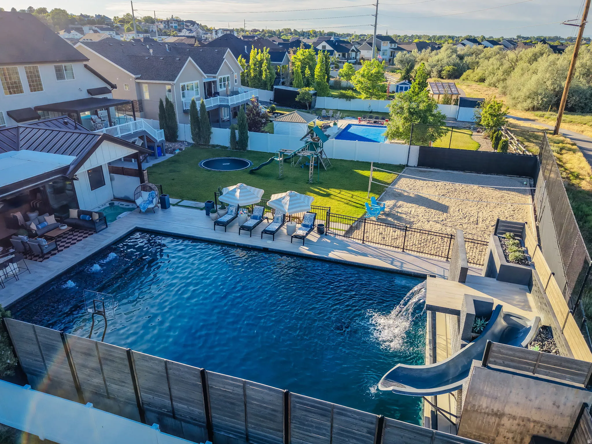 Expansive backyard featuring a large swimming pool with slide and water features, a dedicated sand court, an established lawn, and an inviting covered patio with outdoor seating