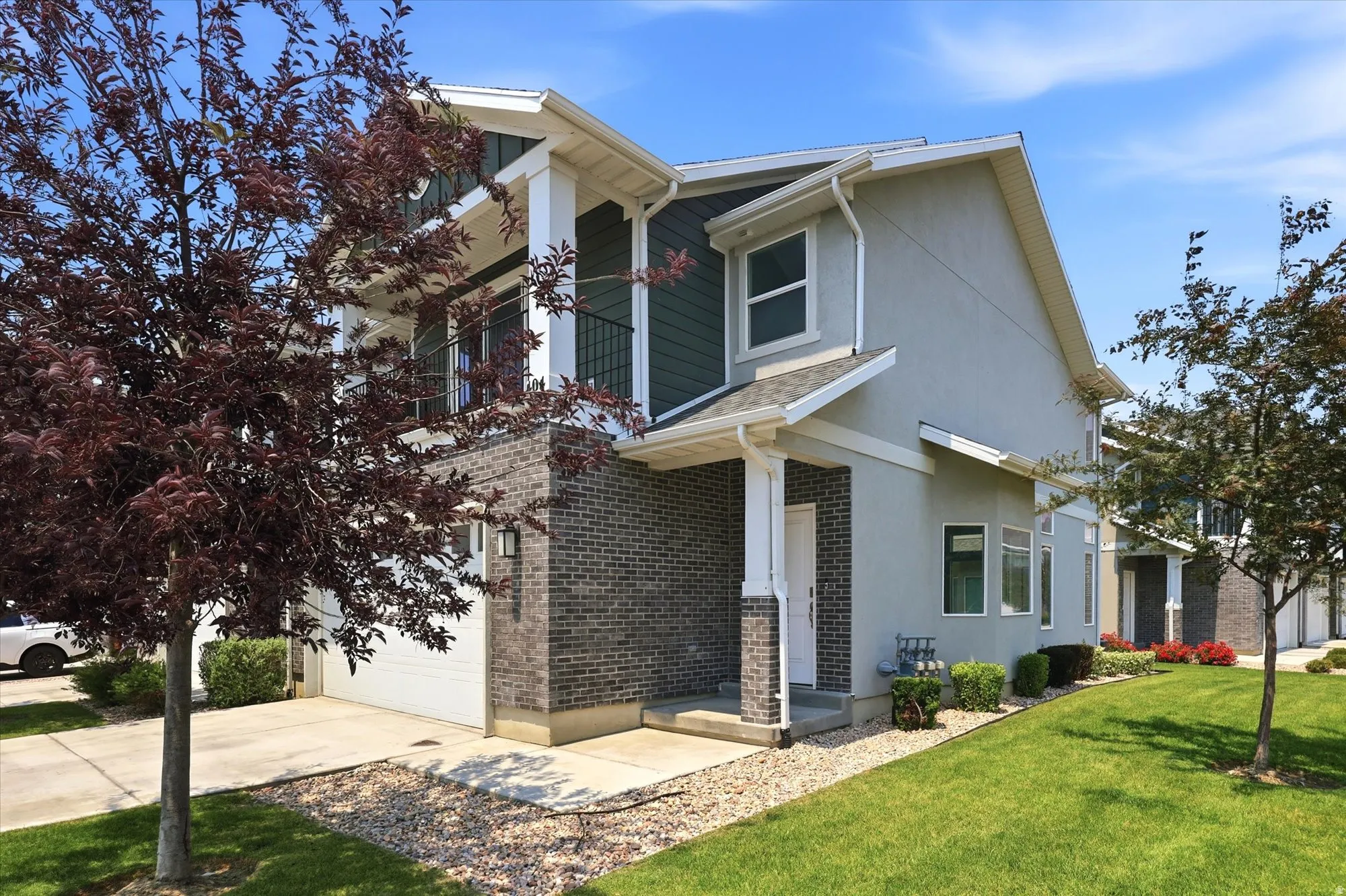Multi-level residential exterior featuring a two-car garage, brick and stucco facade, upper-level balcony with black railing, and mature landscaping with established trees and green lawn