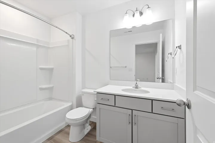 Bathroom featuring a single vanity with quartz-style countertop, white cabinetry with silver hardware, a large wall-mounted mirror, and a white tub and shower combination with an integrated shelving system