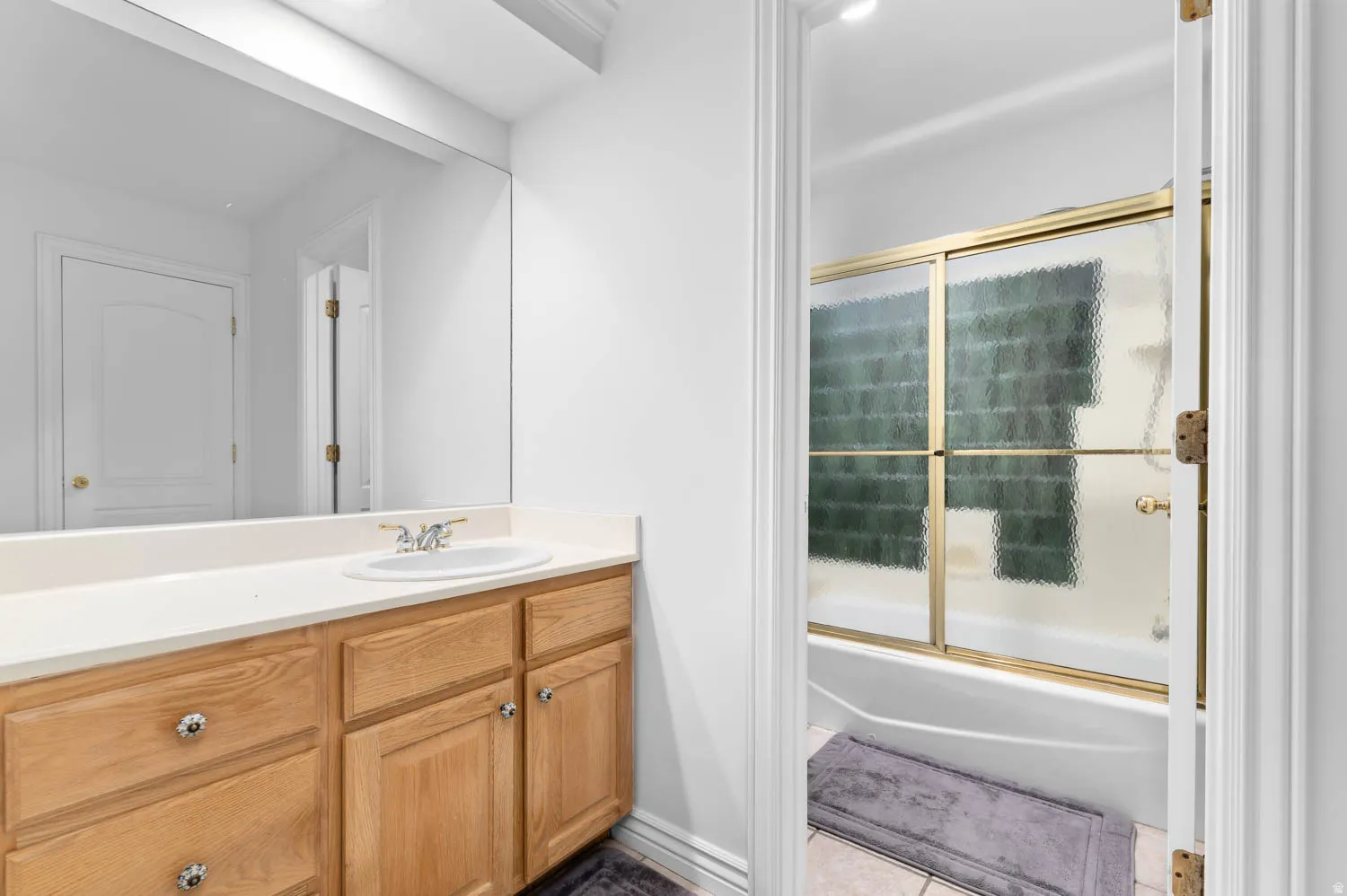 Bathroom featuring a wood-finish vanity with an integrated sink, a large mirror, and an adjacent shower-over-tub with gold-tone framed sliding glass doors