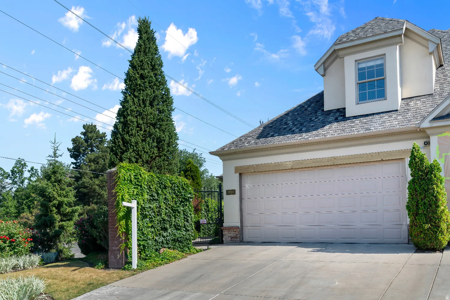 Concrete driveway leading to a two-car garage with a panel door, showcasing a dormer with a multi-pane window, stone facade accents, and surrounding mature landscaping