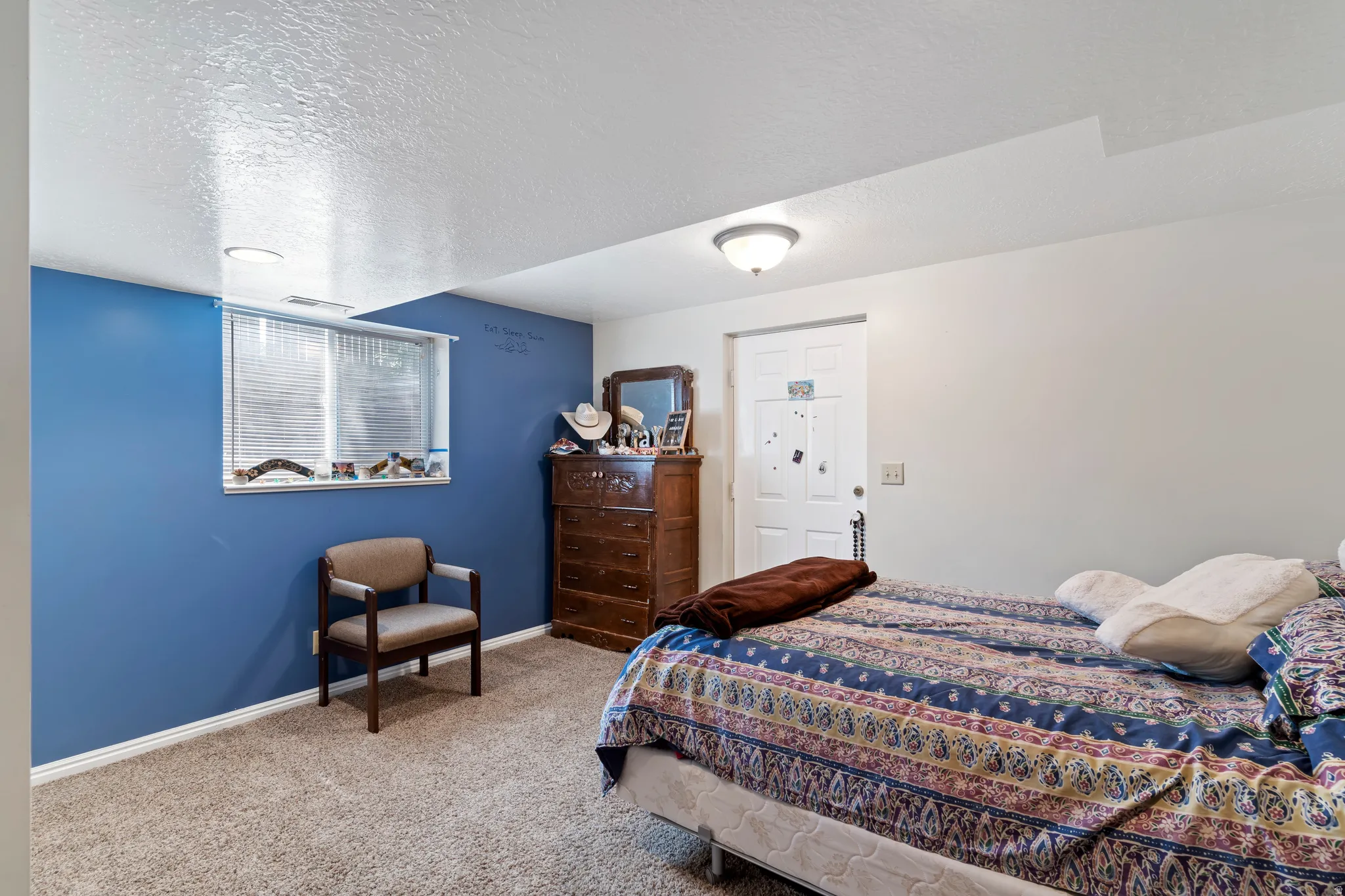 Bedroom featuring blue accent wall, neutral carpeting, a single window with blinds, and a flush mount ceiling light fixture
