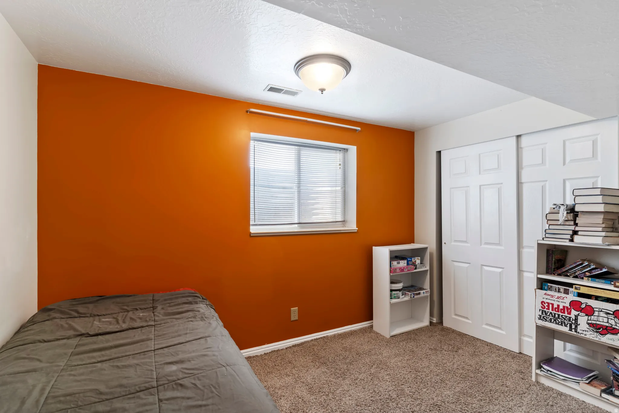 Room featuring orange accent wall, window with blinds, overhead flush mount lighting, carpeted flooring, and a two-panel sliding closet door