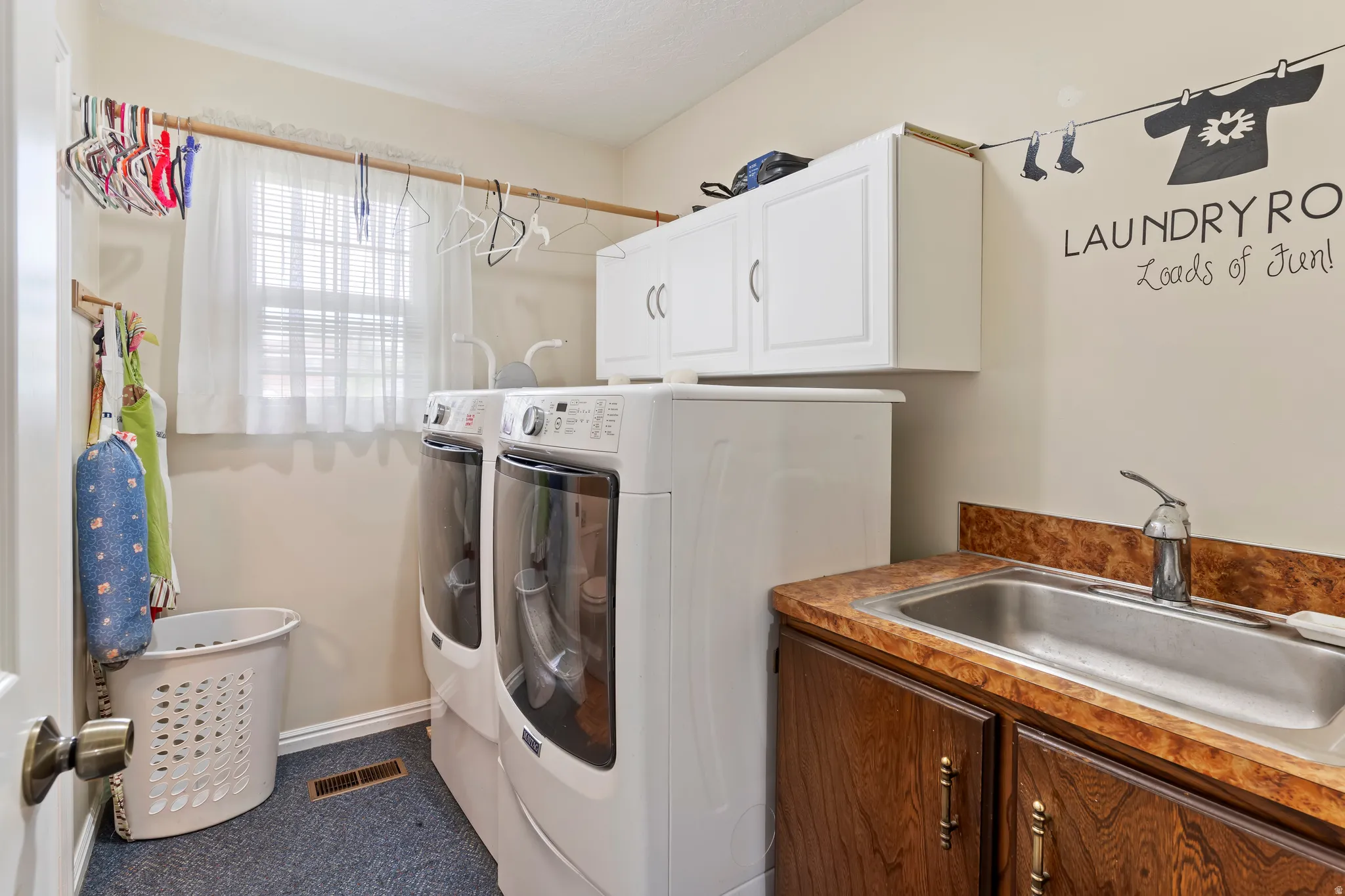 Dedicated laundry room featuring a utility sink with a stainless steel basin, wood-finish base cabinetry, and white overhead storage cabinets