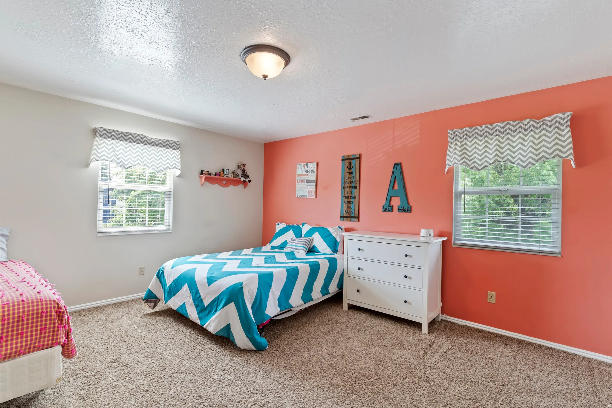 Carpeted bedroom featuring two windows with horizontal blinds, a white dresser with black hardware, and a ceiling-mounted light fixture