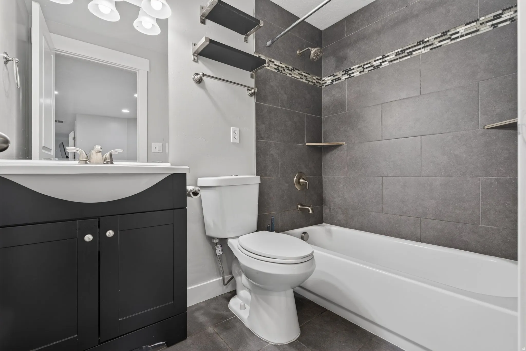 Basement Level full bathroom featuring a dark wood-finish vanity with an integrated sink, brushed nickel hardware, gray floor tiling, and a tiled shower surround with an accent mosaic band