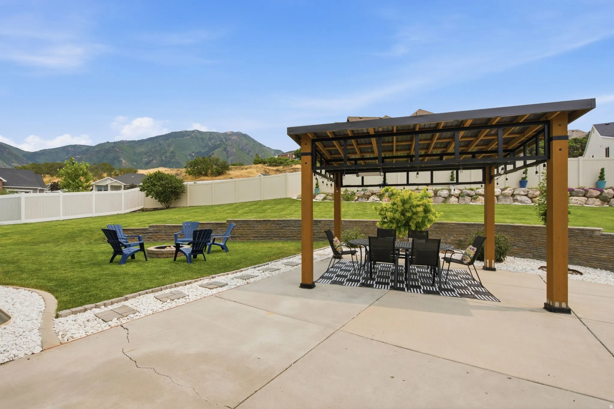 Expansive backyard for outdoor dining and hosting featuring a pergola-covered patio with concrete flooring, a fire pit area, a fully manicured landscape, and a stone retaining wall