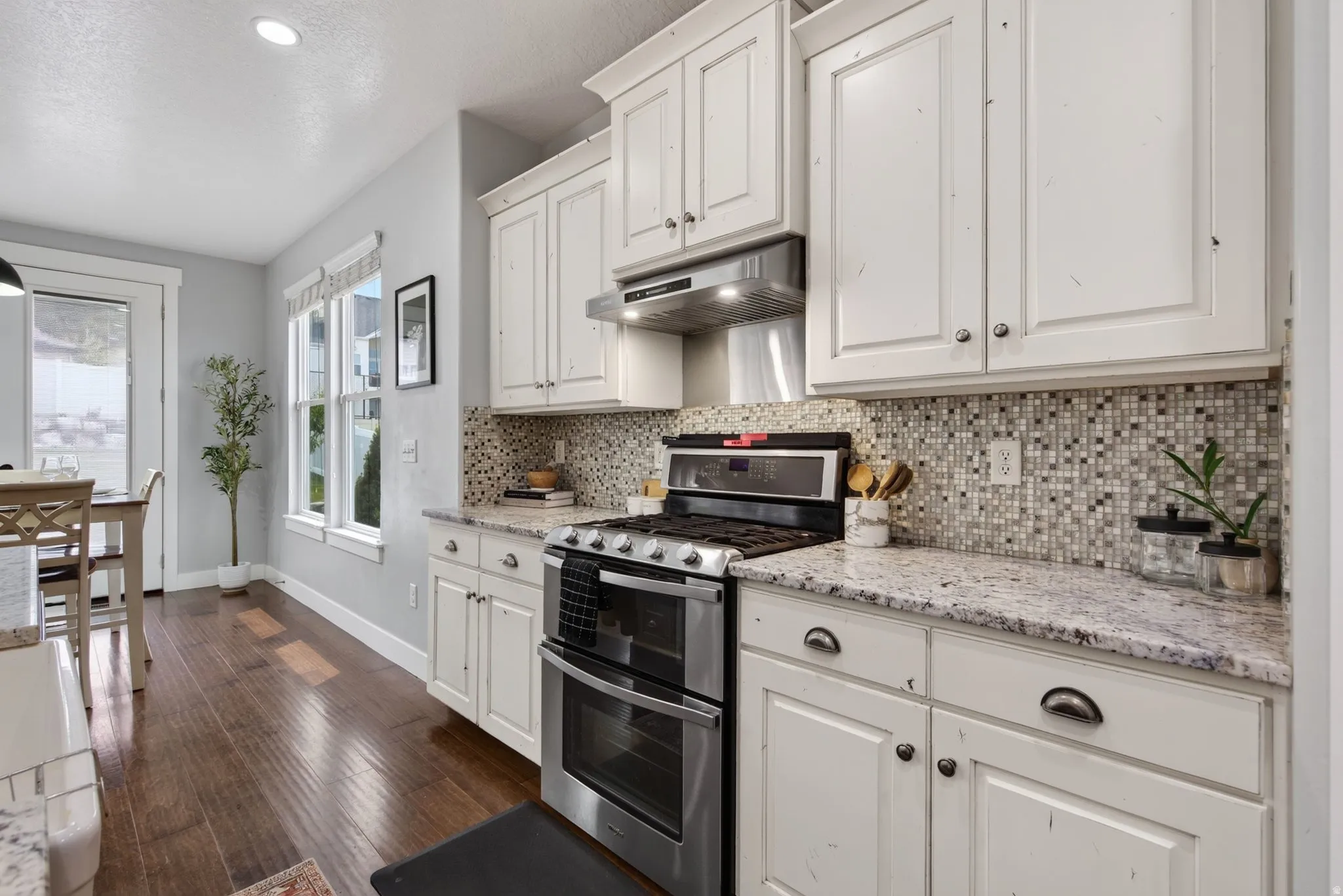 Kitchen view showcasing granite countertops, upgraded range hood vented outside, stainless appliances, tile backsplash, and natural light from the dining area.
