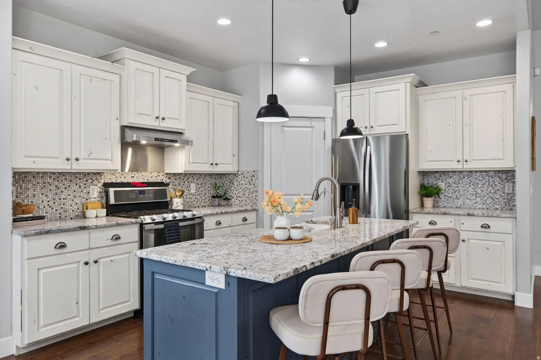 Bright kitchen with granite countertops, large island seating, white cabinetry, stainless appliances, pendant lighting, and an open connection to the living area.