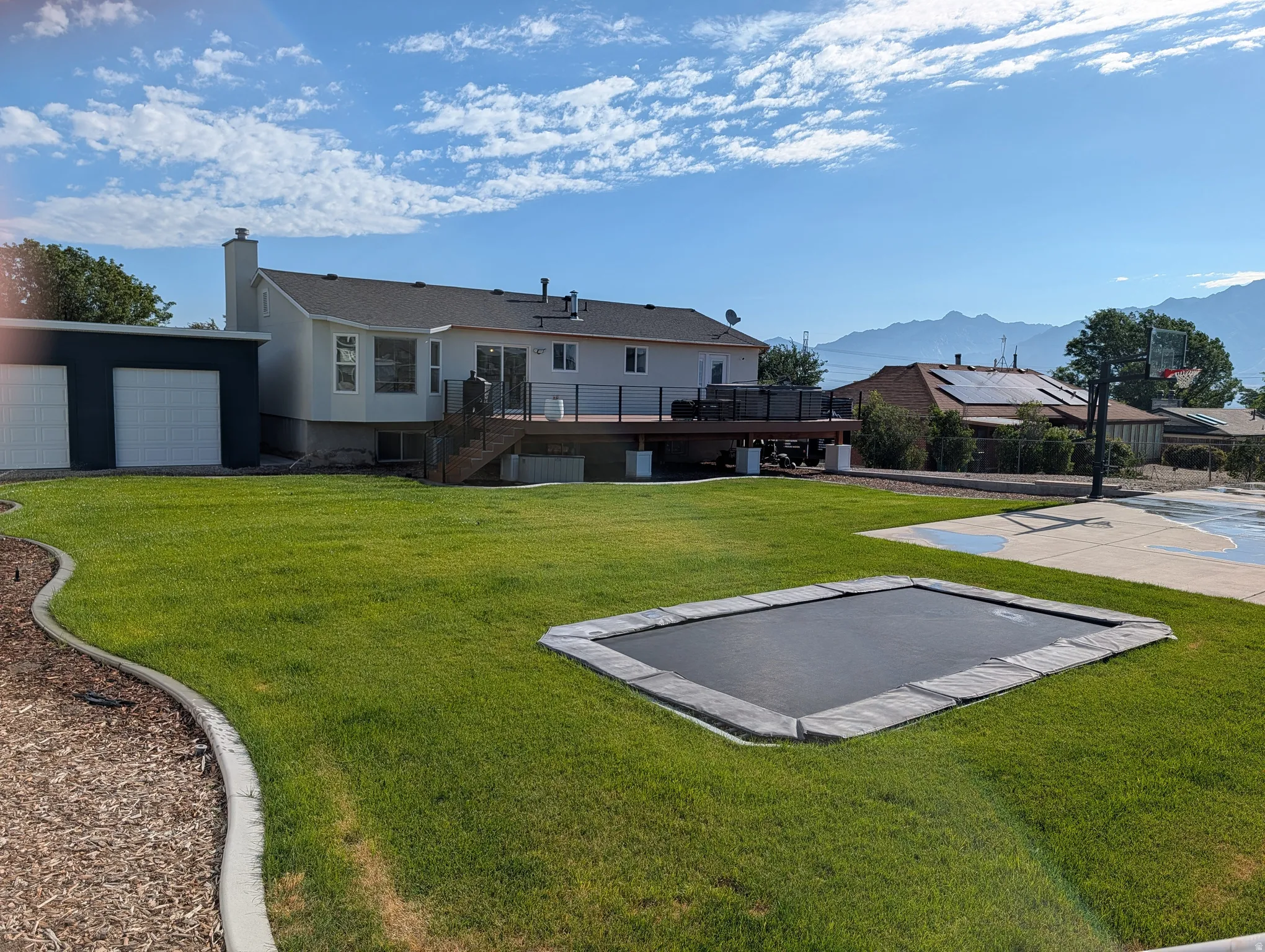 Expansive backyard featuring a manicured lawn, a large wood-finish deck, an in-ground trampoline, and a dedicated sports court with a basketball hoop