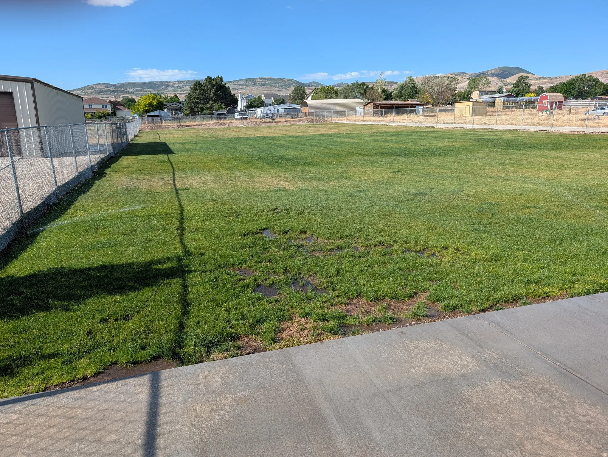 Expansive green space/ball field featuring a chain-link fence, concrete pathway, and distant mountain backdrop