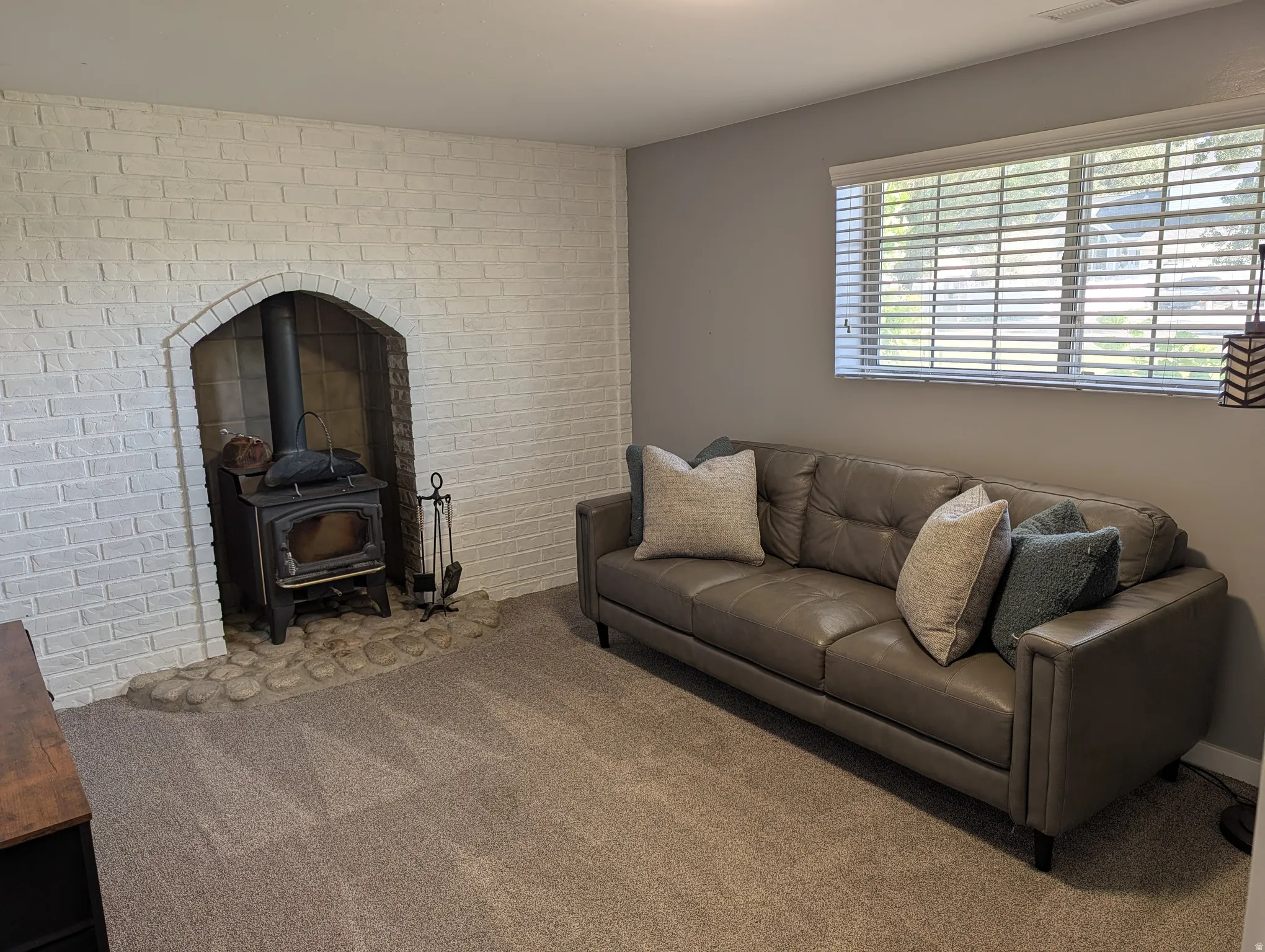 Living area featuring a white-painted brick fireplace with a wood-burning stove, a stone hearth, carpet flooring, and a window with blinds