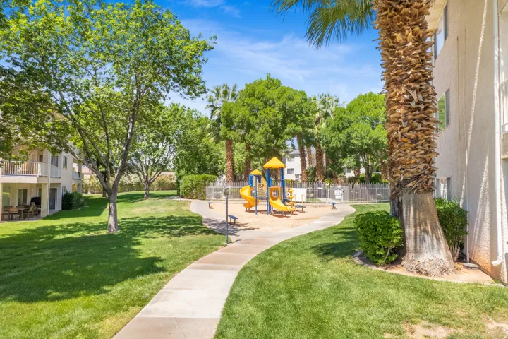 Outdoor recreation area featuring a playground with slides and climbing structures, a concrete walkway, mature palm trees, manicured green lawn, and a light-colored exterior building with a balcony