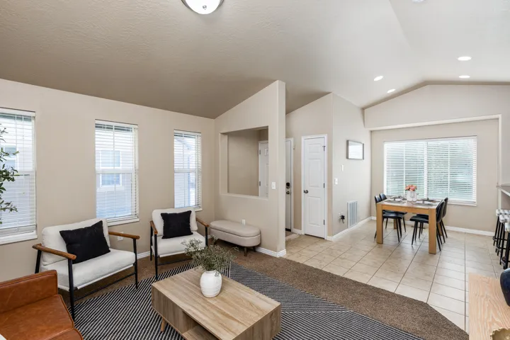Living area with neutral wall tones and multiple windows, featuring vaulted ceilings and a clear sightline to the dining area with tiled flooring