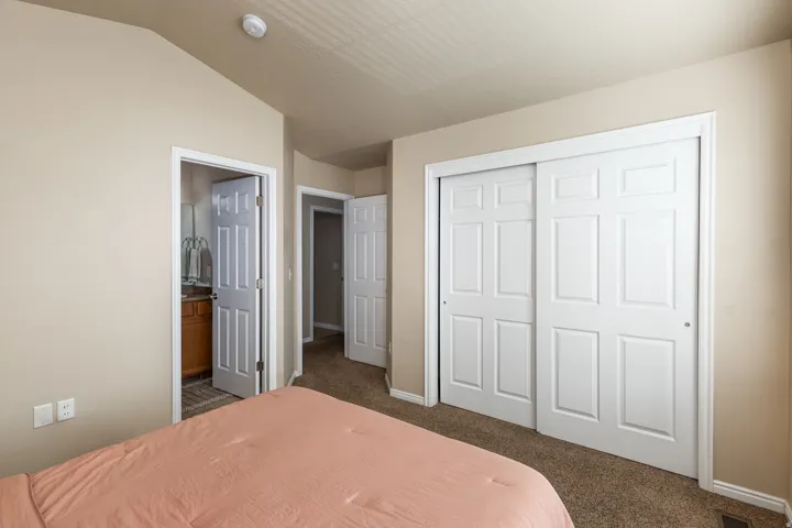 Vaulted ceiling room with neutral paint, carpet flooring, a white sliding closet door, and a six-panel door entry to a bathroom featuring a wooden vanity and mirror