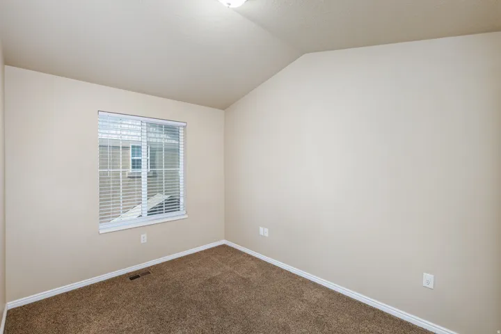 Neutral-toned room featuring a single window with horizontal blinds, textured carpeting, white baseboards, and a sloped ceiling
