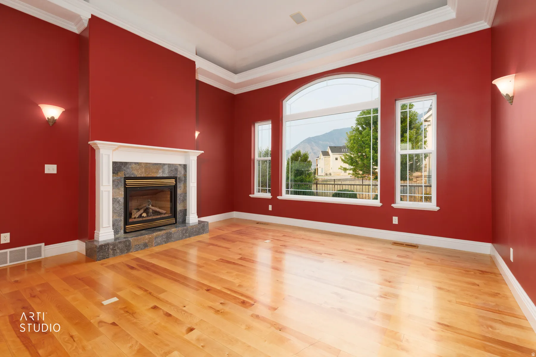 Spacious room featuring hardwood flooring, a gas fireplace with a stone surround and white mantel, large arched windows, and crown molding