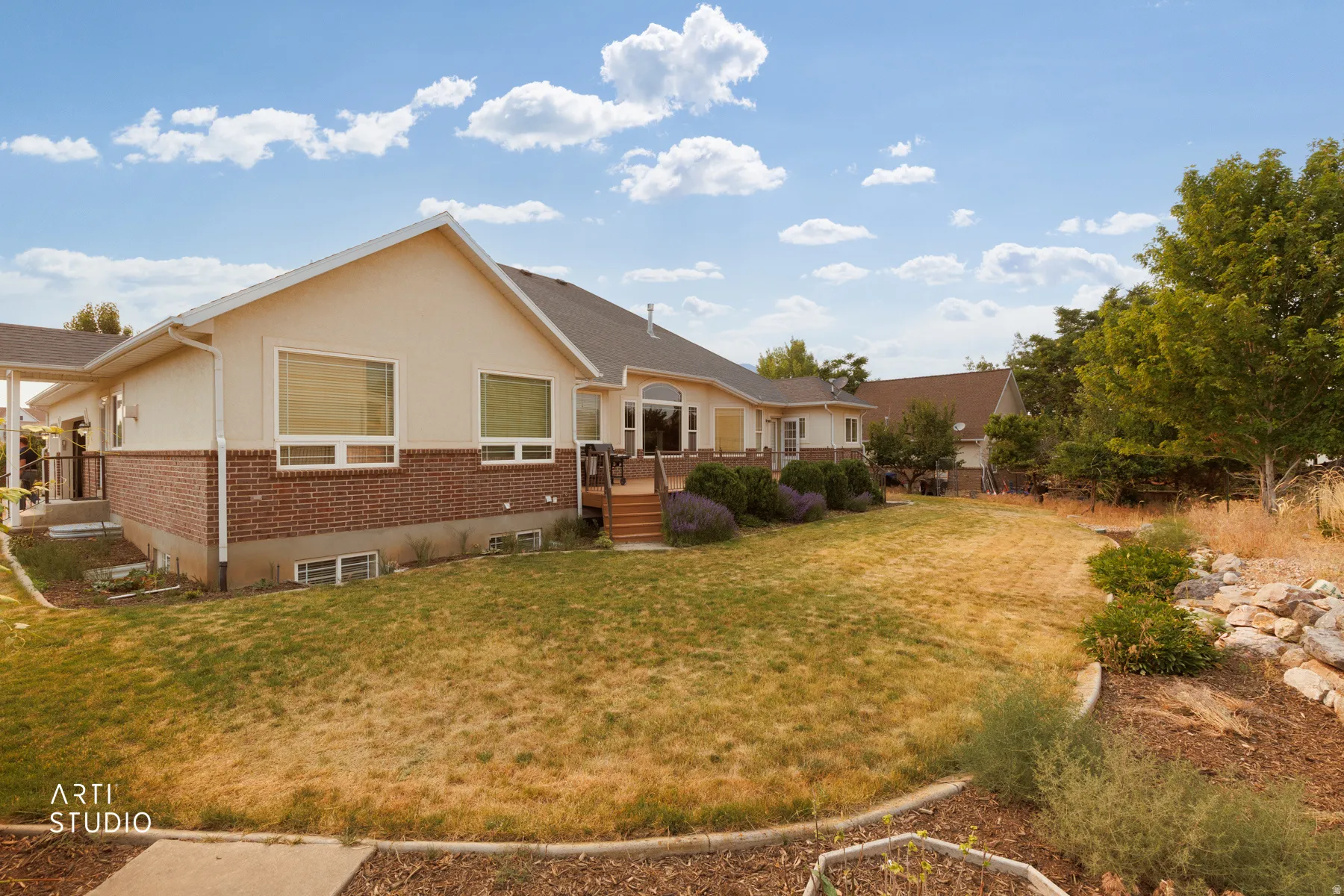 Rear exterior featuring a brick and stucco facade, multiple windows, a wooden deck with stairs, a patio, and mature landscaping
