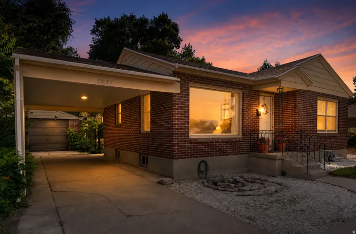 Brick exterior featuring a covered carport with recessed lighting, a front porch with wrought iron railings, a prominent picture window, and a gravel-covered front yard with a rock feature