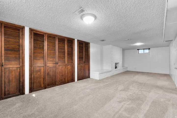 Spacious carpeted room featuring a white painted brick fireplace with hearth seating, a window providing natural light, and a wall of wood-louvered bi-fold doors