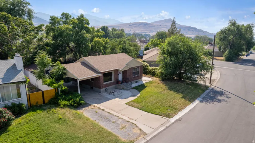 Brick exterior home featuring a covered carport, mature trees, a landscaped front yard with a circular stone patio, and a corner lot position