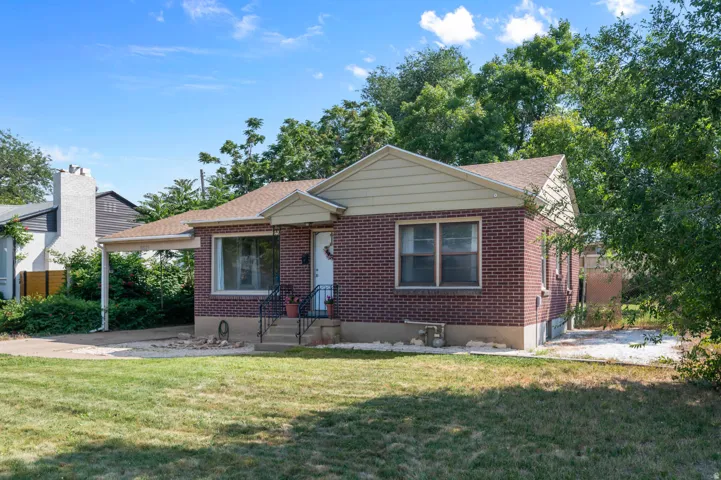 Brick exterior home featuring a covered carport, gabled roofline, front entry porch with wrought iron railing, and a sizable lawn area