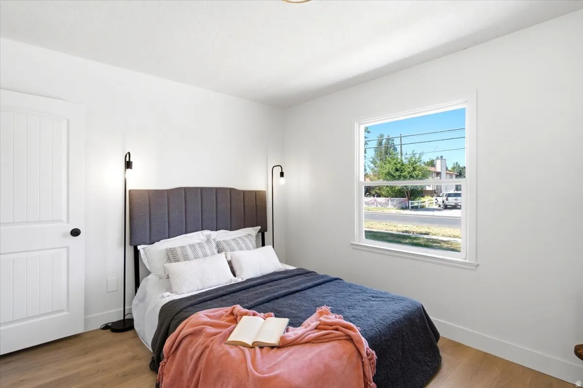 Bright bedroom featuring a white door with black hardware, wood-finish flooring, and a large window with white trim