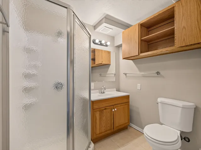 Bathroom featuring a glass-enclosed shower, light wood-finish cabinetry, a white vanity with integrated sink, and neutral tile flooring
