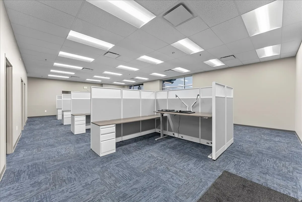 Open office space featuring patterned blue carpet, grid ceiling with recessed lighting, light-toned walls, and multiple workstation cubicles