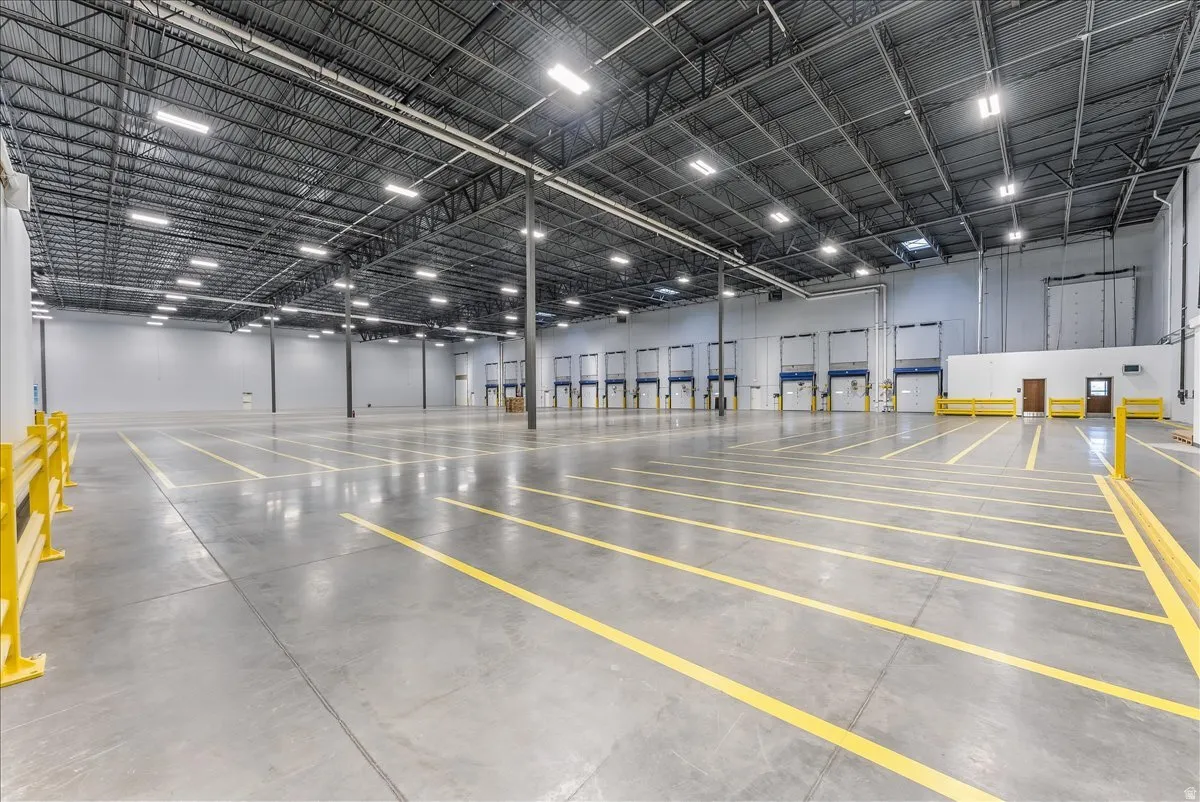 Spacious industrial warehouse featuring multiple overhead dock doors, high ceilings with exposed structural elements, abundant overhead lighting, polished concrete flooring, and prominent yellow safety railings