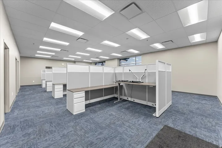Open office space featuring patterned blue carpet, grid ceiling with recessed lighting, light-toned walls, and multiple workstation cubicles