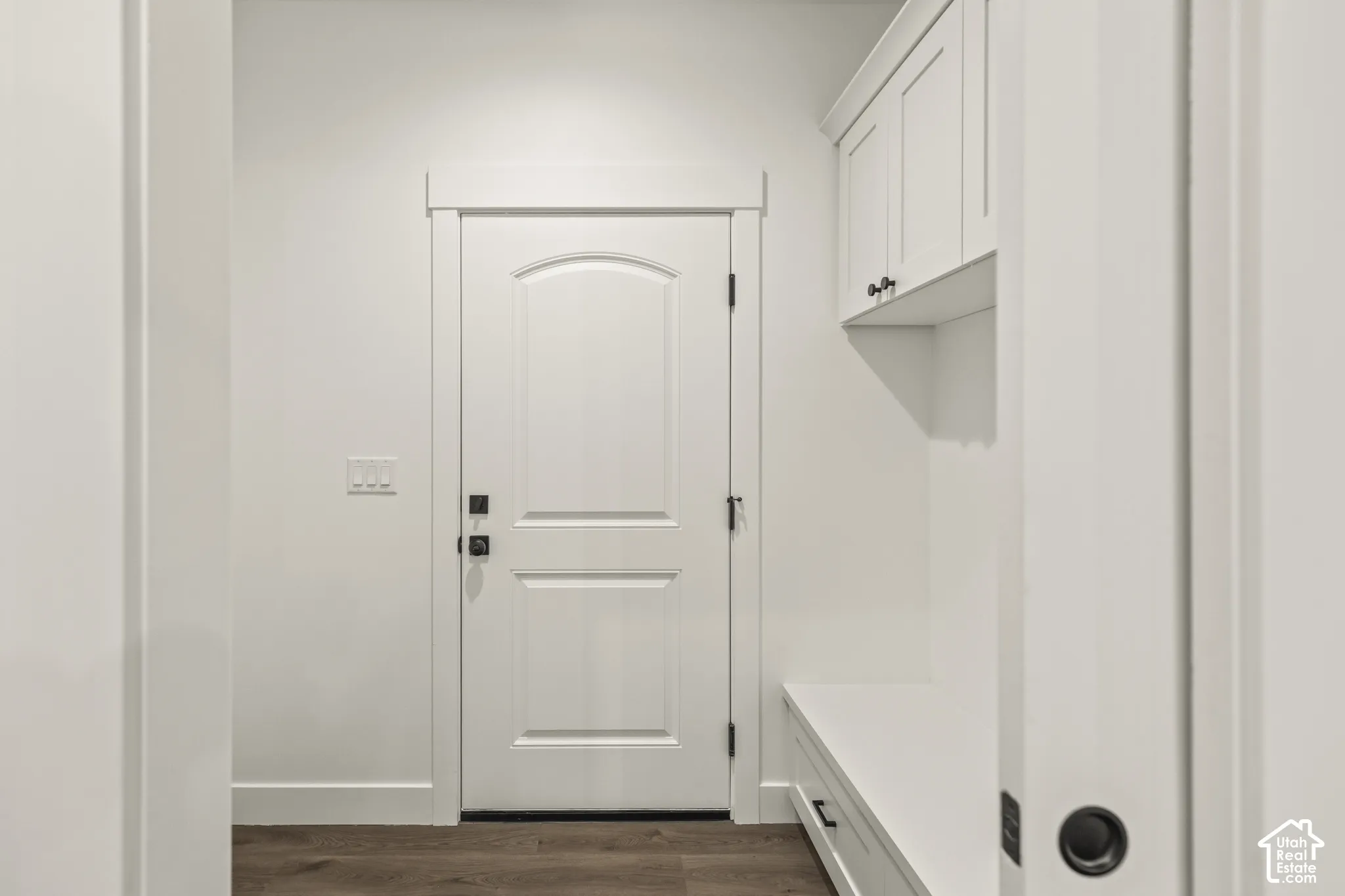 Entryway featuring wood-finish flooring, white paneled door with dark hardware, built-in bench, and overhead cabinetry