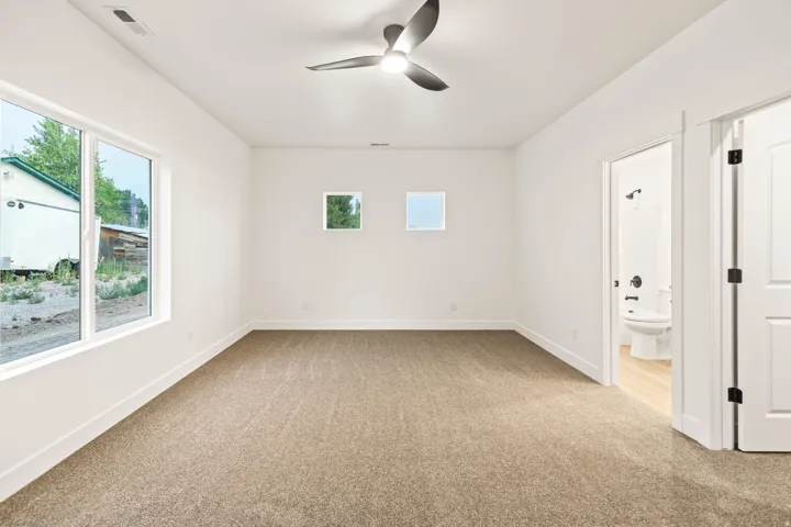 Spacious room featuring neutral carpeting, a modern ceiling fan, multiple windows for ample natural light, and an attached bathroom with a white toilet and dark fixtures