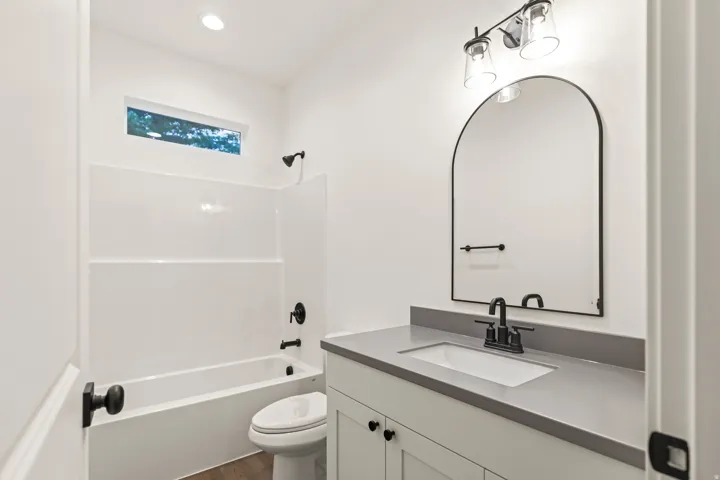 Bathroom featuring a white vanity with a gray countertop, an arched black-framed mirror, a tub-shower combination with a white surround, and dark bronze fixtures