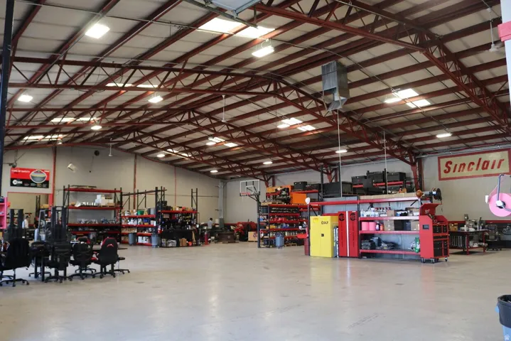 Expansive warehouse space featuring a high-pitched metal roof with exposed red trusses, numerous fluorescent light fixtures, and multiple skylights
