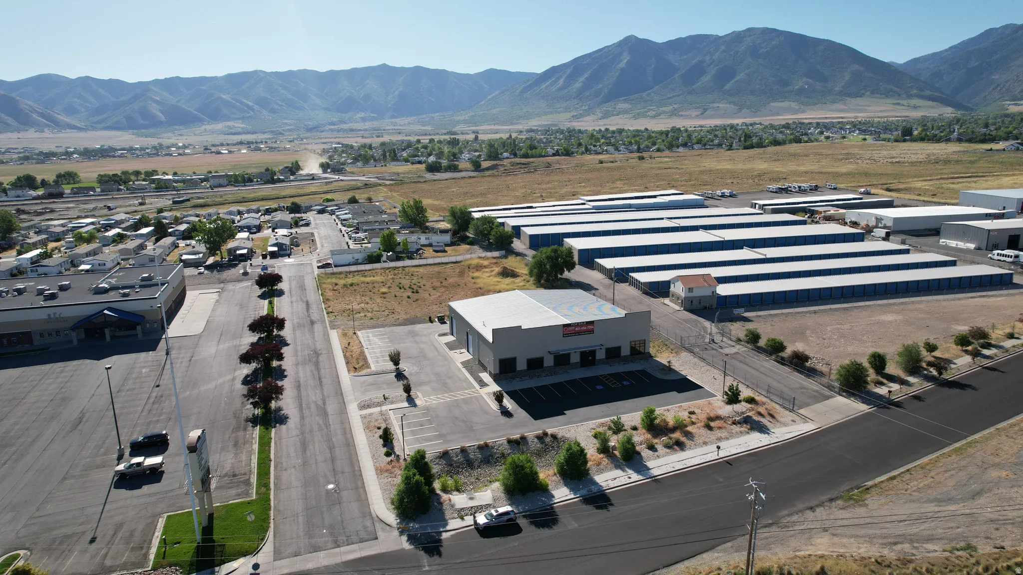 Commercial property featuring a gray facade and flat roof, ample paved parking, established landscaping with trees and shrubs, and a clear, sunny mountain backdrop