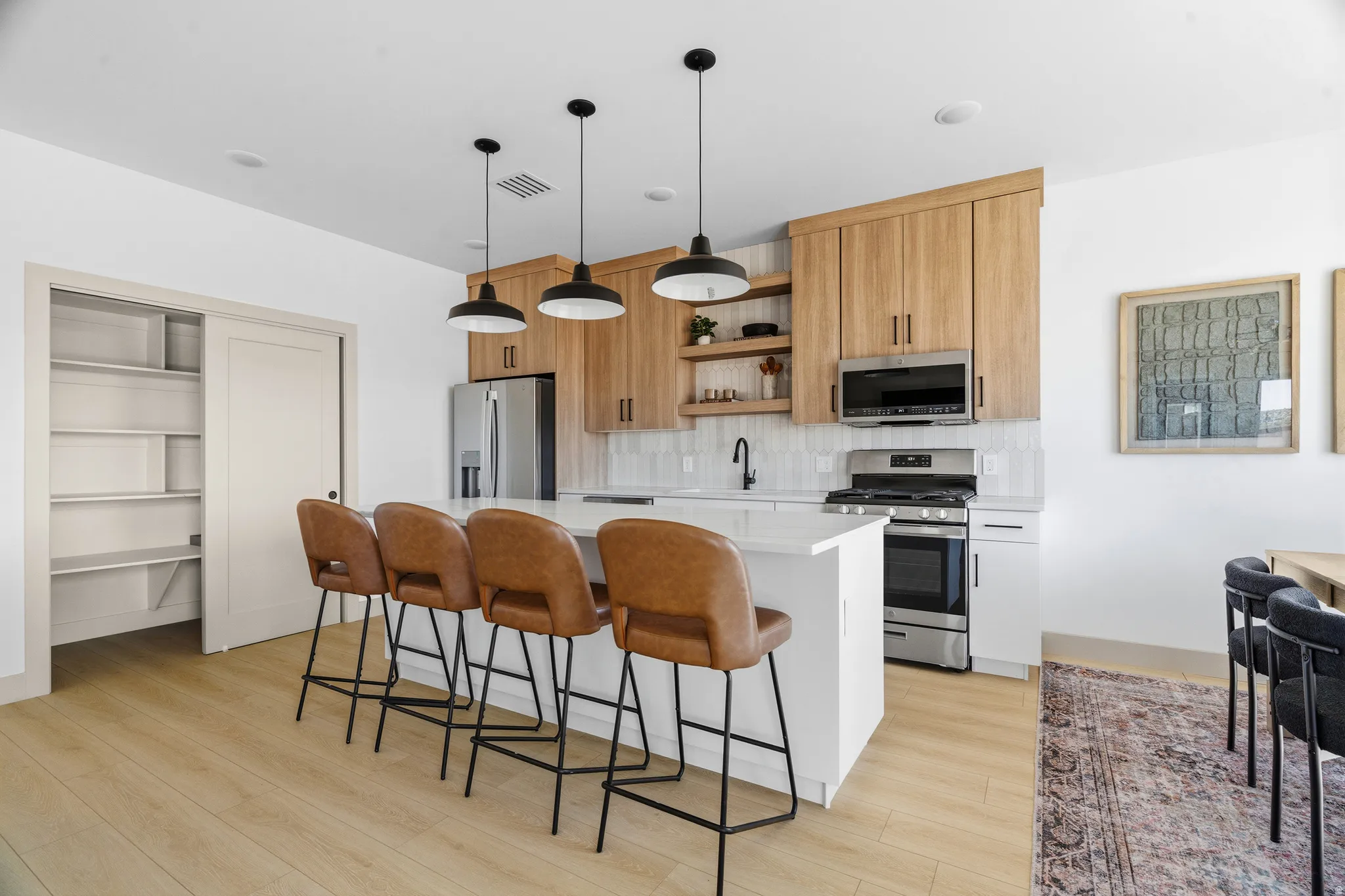 Modern kitchen featuring light wood-finish flooring, a white countertop island with pendant lighting, stainless steel appliances, and a tiled backsplash