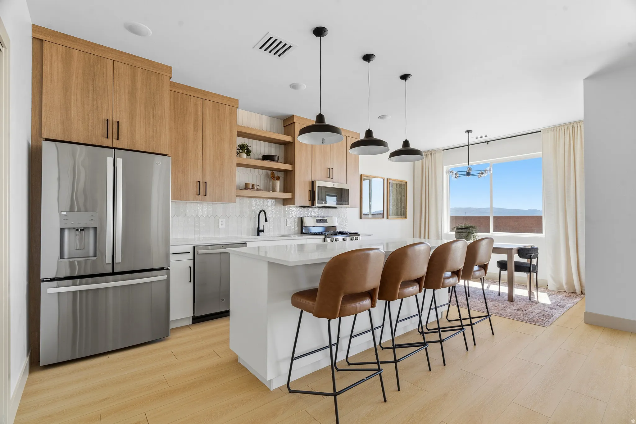 Open-concept kitchen with wood-finish flooring, featuring a stainless steel refrigerator, white tile backsplash, and a central island with countertop seating
