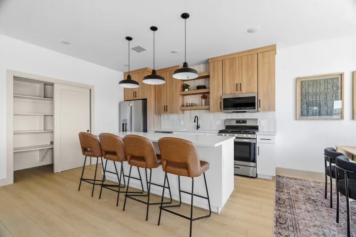 Modern kitchen featuring light wood-finish flooring, a white countertop island with pendant lighting, stainless steel appliances, and a tiled backsplash