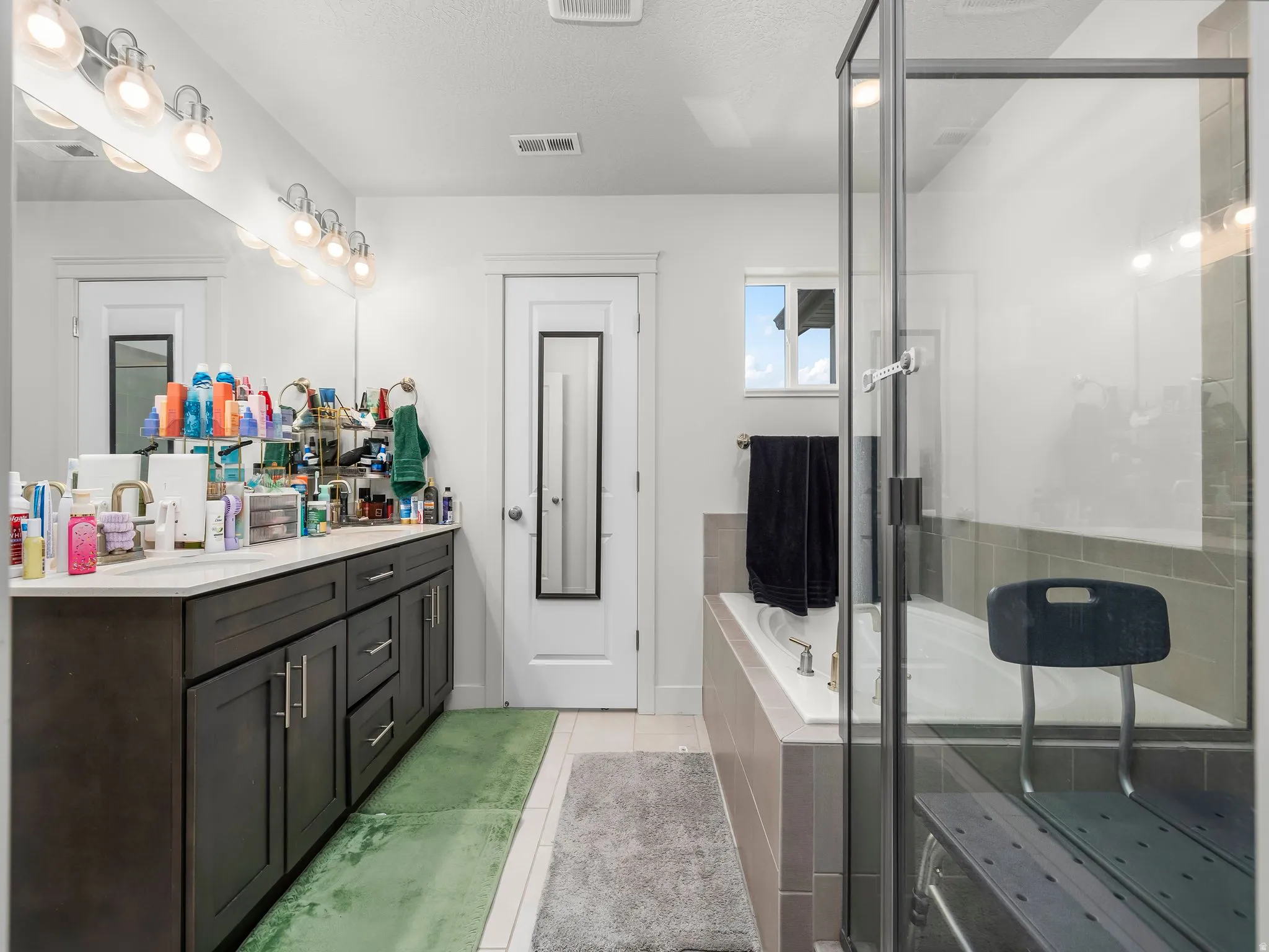 Bathroom featuring a dual vanity with dark wood-finish cabinetry, a large frameless mirror, a built-in soaking tub with tiled surround, and a glass-enclosed shower