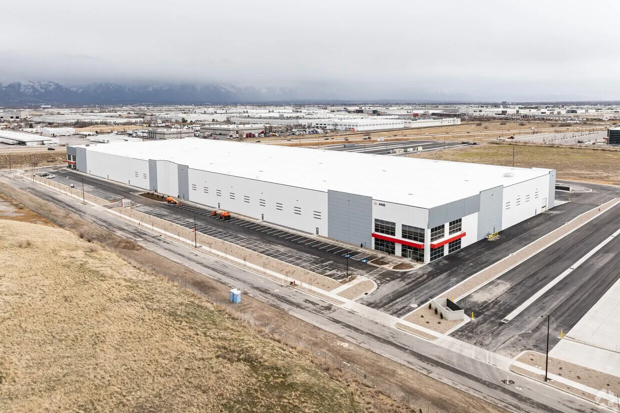 Expansive industrial building featuring a white and gray exterior, extensive parking, multiple loading docks, snow-capped mountain backdrop, and surrounding flat terrain