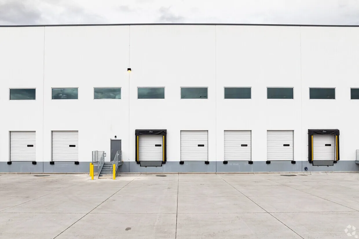 Industrial building exterior featuring a white facade, multiple dock-high doors, upper-level windows, and a concrete loading area