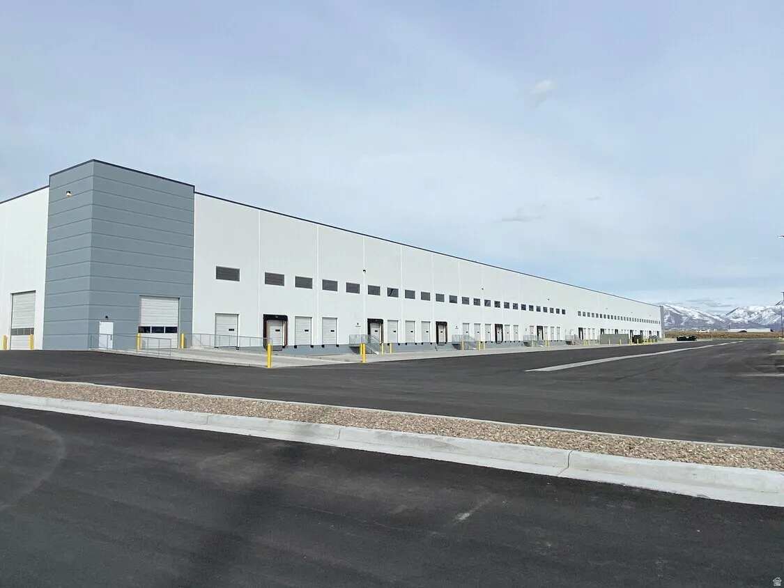 Industrial facility featuring a white and gray exterior, numerous dock-height loading doors, multiple ground-level roll-up doors, an expansive paved lot, and a mountain backdrop