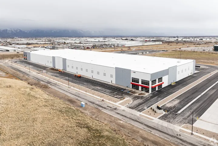 Expansive industrial building featuring a white and gray exterior, extensive parking, multiple loading docks, snow-capped mountain backdrop, and surrounding flat terrain